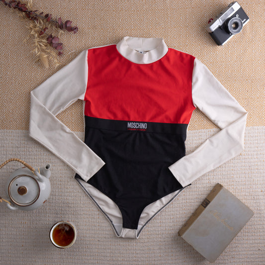 Vintage Moschino "Under Where?" Y2K Long Sleeve Bodysuit – red, black and white colorblock stretch fabric, mock turtleneck, long white sleeves, red chest panel, black lower body with embroidered Moschino logo band, snap crotch, 90s streetwear, flat lay editorial shot