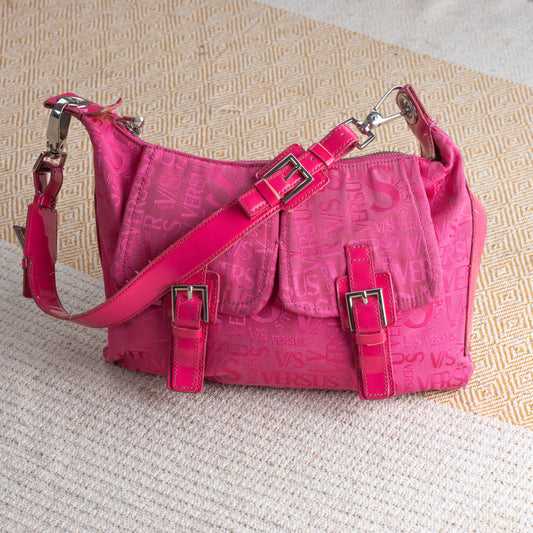 Vintage Versus Versace Y2K Pink Monogram Shoulder Bag – hot pink embossed nylon with all-over tonal Versus logo lettering, patent leather trim and adjustable crossbody strap, two front flap pockets with silver buckle hardware, top zip closure, silver carabiner clips, Barbiecore McBling aesthetic, product shot on textured surface
