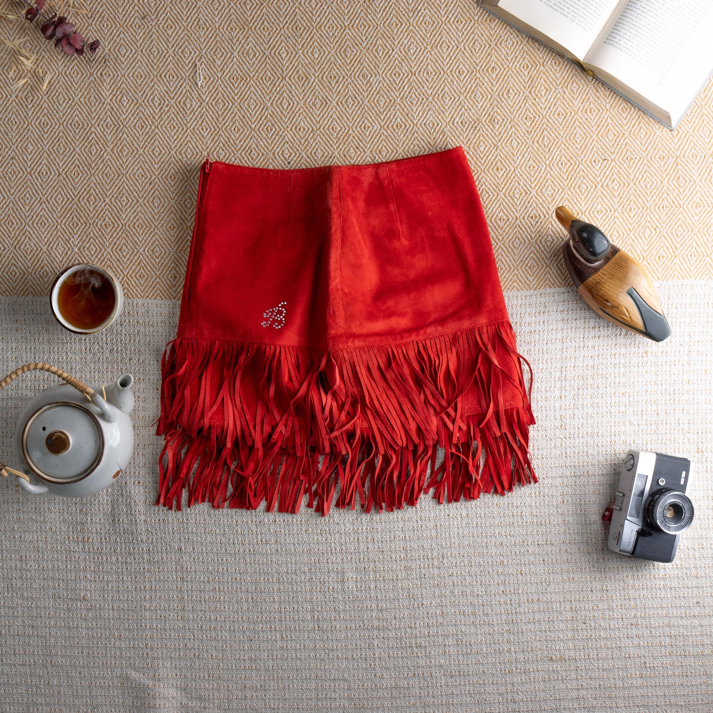Vintage Blumarine Folies Y2K Red Suede Fringe Mini Skirt – vibrant red genuine leather suede, high waist, A-line silhouette with dramatic double-layer fringe hem, rhinestone crystal "B" logo detail at hip, western glam aesthetic, flat lay editorial shot
