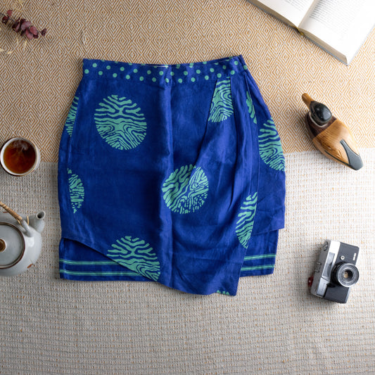 Vintage Versus Versace 90s Linen Mini Skirt – royal blue linen with bold oversized green zebra-print medallion pattern, green polka dot waistband, wrap-style asymmetric front panel, green stripe hem border, Italian summer luxury, flat lay editorial shot