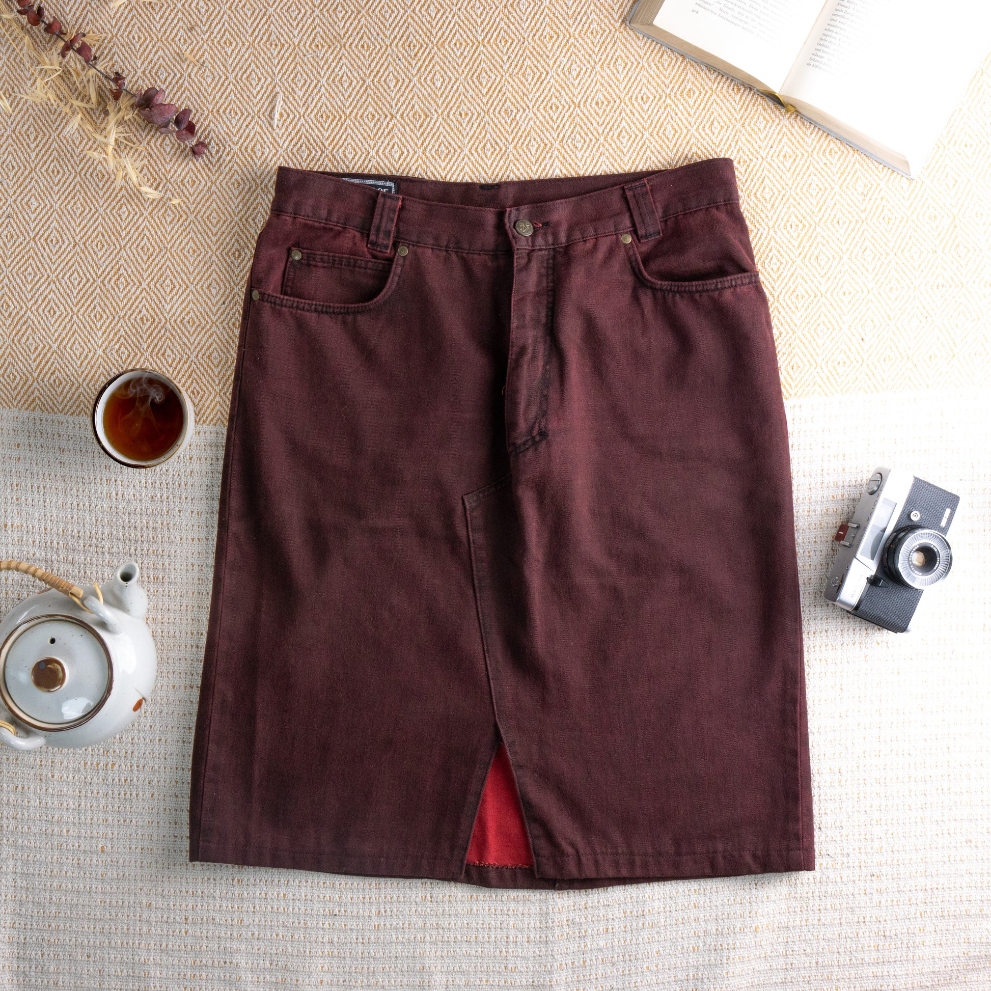 Reworked Versace Denim Midi Skirt – burgundy wine overdyed denim, mid rise with five-pocket construction and silver rivet hardware, pencil silhouette with deconstructed raw hem front slit revealing red lining, upcycled avant-garde grunge rework, flat lay editorial shot