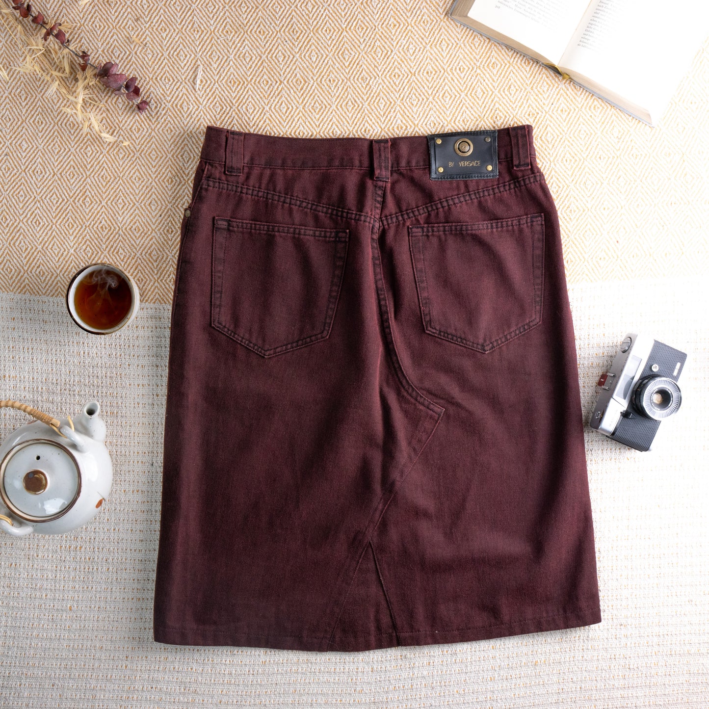 back view Reworked Versace Denim Midi Skirt – burgundy wine overdyed denim, mid rise with five-pocket construction and silver rivet hardware, pencil silhouette with deconstructed raw hem front slit revealing red lining, upcycled avant-garde grunge rework, flat lay editorial shot
