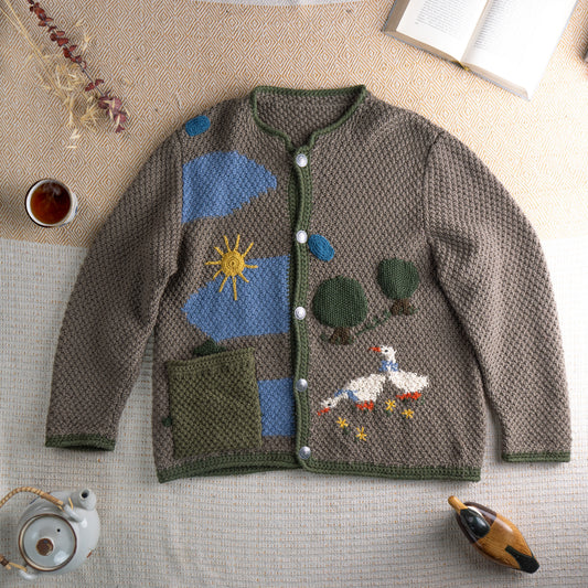 Vintage Italian Alpine Udine Hand Knit Oversized Cardigan – taupe brown chunky knit with green trim, intarsia pastoral scene featuring blue sky, yellow sun, green trees and white goose, silver coin buttons, two patch pockets, round neckline, long sleeves, artisan folk craft, flat lay editorial shot