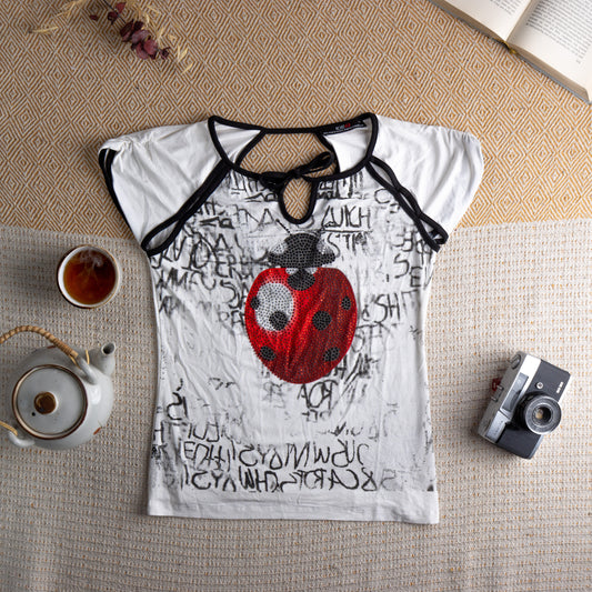 ICE B Iceberg Deadstock Y2K Rhinestone Ladybug T-Shirt – white viscose with all-over black graffiti letter print, oversized red and black rhinestone ladybug appliqué at centre front, black contrast trim, cut-out keyhole neckline with strappy detail, short cap sleeves, McBling 2000s, flat lay editorial shot
