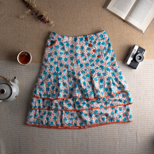 Cacharel Archival Deadstock Tiered Ruffle Skirt – white silk-cotton blend with all-over turquoise blue abstract daisy print and red dot scatter, drop waist with three red button front closure, small side pocket with orange trim, double tiered ruffle hem with orange grosgrain ribbon trim, coquette French chic quiet luxury, flat lay editorial shot