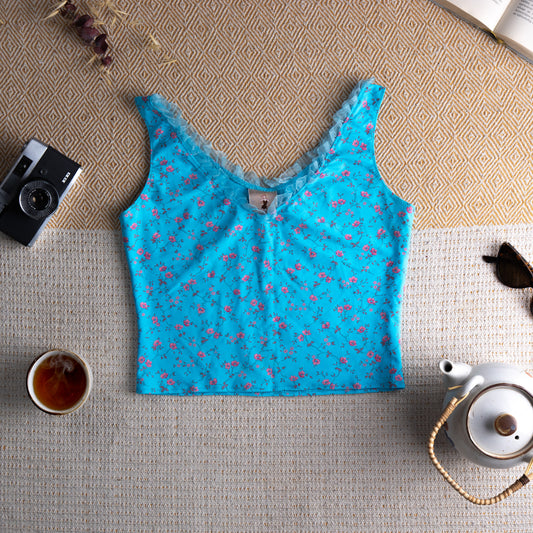 Blumarine Y2K Vintage Camisole Crop Top – turquoise blue stretch fabric with all-over pink ditsy rose floral print, V-neckline with ruffled lace trim, wide shoulder straps with lace edging, fitted cropped silhouette, Blumarine logo label at centre front, McBling coquette 2000s aesthetic, flat lay editorial shot