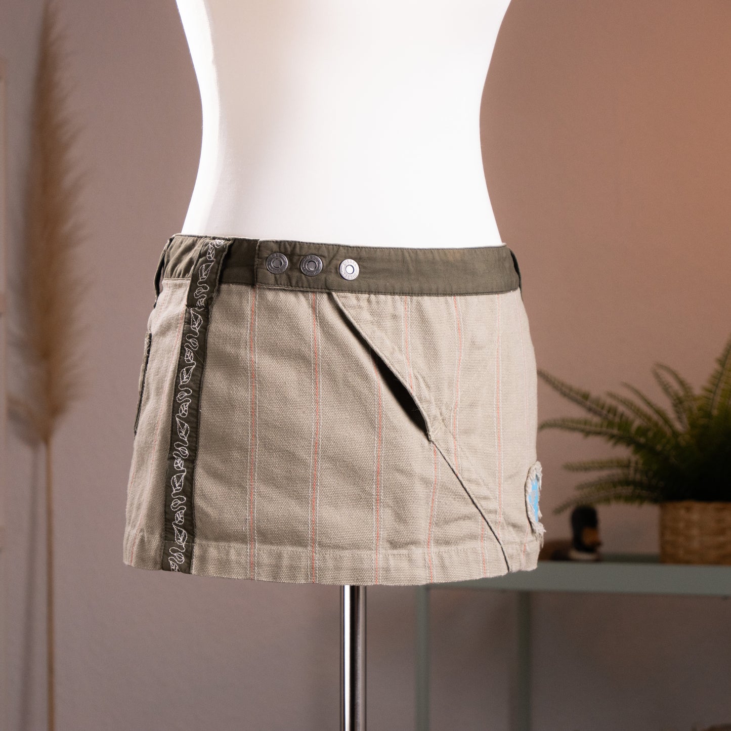 Three-quarter view of the archival Diesel khaki canvas wrap mini skirt, highlighting the architectural diagonal slash pocket, the low-rise twill waistband, and the industrial metallic snap buttons.
