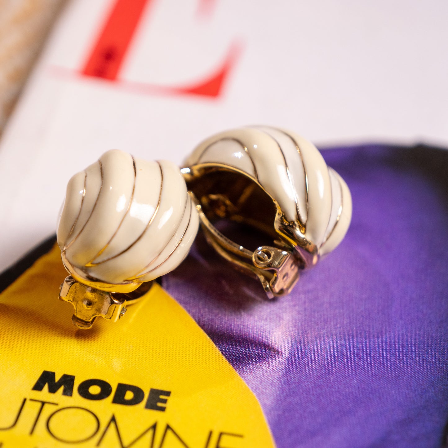 Detailed view of the archival 1980s Nina Ricci clip earrings, demonstrating a 1-of-1 factory error with one earring featuring finished white enamel in the recessed channels and the other showing completely unfilled raw base grooves.

